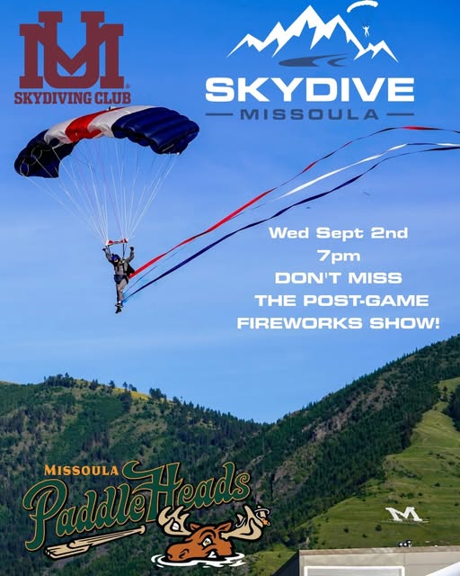 Skydiver under a red white and blue canopy trailing a banner over Missoula, promoting a demo jump at a Paddleheads game